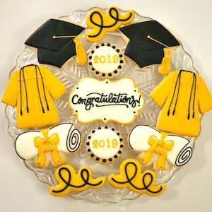 May include: A set of nine graduation-themed sugar cookies decorated with yellow and black icing. The cookies feature a graduation cap, a diploma, a gown, and the text "Congratulations!" and "2019".