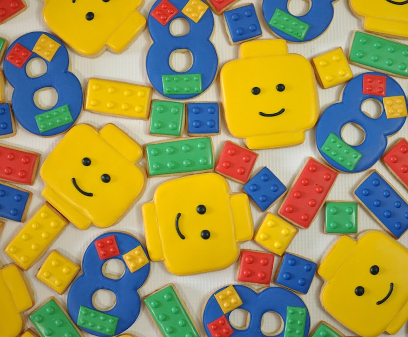 May include: Assortment of colorful, decorated sugar cookies shaped like Lego bricks and numbers. The cookies feature various designs, including smiley faces, numbers, and Lego brick patterns.