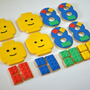 May include: A set of colorful Lego-themed sugar cookies, including four yellow Lego head cookies, three blue number eight cookies decorated with smaller Lego brick designs, and six smaller Lego brick cookies in red, blue, and yellow.