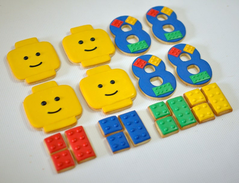 May include: A set of colorful Lego-themed sugar cookies, including four yellow Lego head cookies, three blue number eight cookies decorated with smaller Lego brick designs, and six smaller Lego brick cookies in red, blue, and yellow.