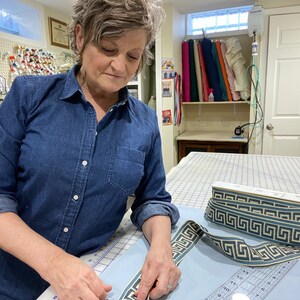 Op de afbeelding: Een persoon werkt aan een naaiproject en meet een blauw en wit gepatroneerd lint met een liniaal. Het lint ligt op een lichtblauwe stof, en er is een witte spoel lint op de achtergrond.