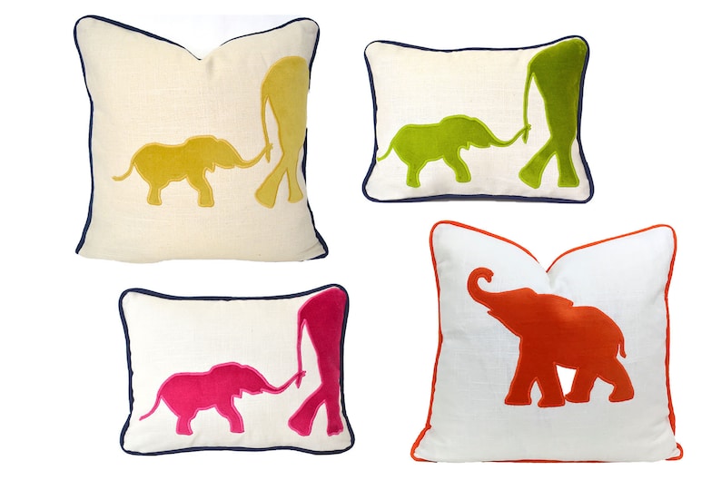 Op de afbeelding: Vier decoratieve kussens met geborduurde olifantensilhouetten. De kussens zijn wit met een marineblauwe rand. De olifanten zijn in verschillende kleuren: geel, groen, roze en oranje.