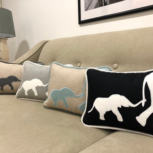 Op de afbeelding: Vier decoratieve kussens met geborduurde olifantensilhouetten. De kussens zijn in een rij op een lichtbruine bank gerangschikt. De olifanten zijn in verschillende kleuren, waaronder grijs, wit, blauw en zwart.