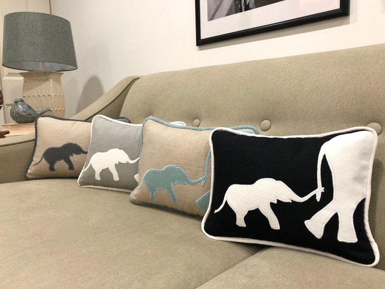 Op de afbeelding: Vier decoratieve kussens met geborduurde olifantensilhouetten. De kussens zijn in een rij op een lichtbruine bank gerangschikt. De olifanten zijn in verschillende kleuren, waaronder grijs, wit, blauw en zwart.