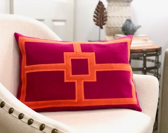 May include: A rectangular magenta pillow with an orange geometric design. The pillow is placed on a cream-colored chair. The geometric pattern is a series of connected squares and rectangles.