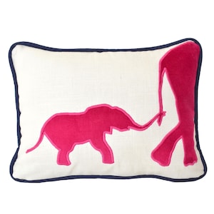 Op de afbeelding: Een wit kussen met een donkerblauwe rand heeft een roze geborduurd ontwerp van twee olifanten, een groter dan de andere, waarbij de slurf van de kleinere olifant het been van de grotere raakt.