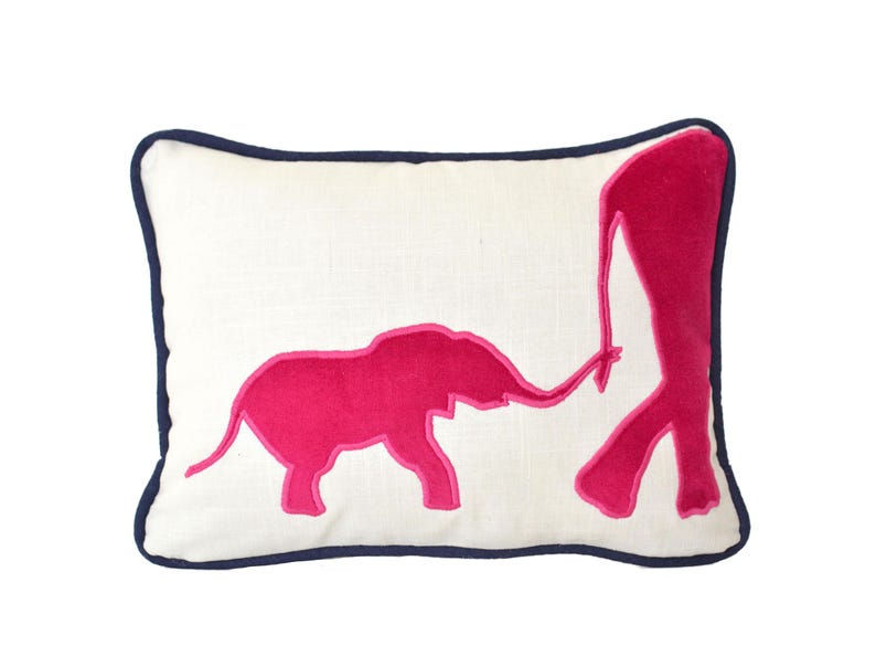 Op de afbeelding: Een wit kussen met een donkerblauwe rand heeft een roze geborduurd ontwerp van twee olifanten, een groter dan de andere, waarbij de slurf van de kleinere olifant het been van de grotere raakt.