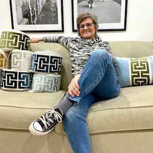 May include: A woman sits on a beige couch with a stack of decorative trim in various colors and patterns. The trim features a geometric design. The woman is wearing blue jeans, a black and white striped sweater, and black sneakers.