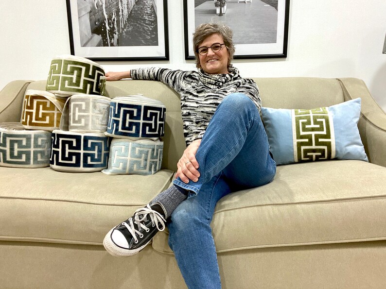 May include: A woman sits on a beige couch with a stack of decorative trim in various colors and patterns. The trim features a geometric design. The woman is wearing blue jeans, a black and white striped sweater, and black sneakers.