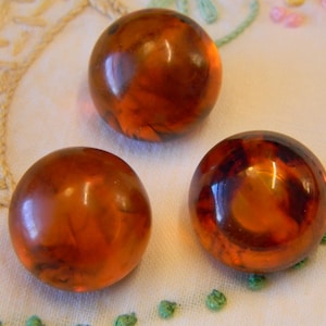 May include: Three translucent, amber-coloured, round beads with a marbled appearance. The beads are arranged on a white surface with embroidered details and small green embellishments.