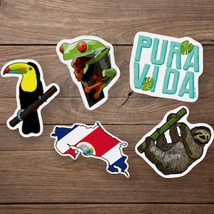 Peut inclure: Ensemble d'autocollants représentant la faune et des phrases du Costa Rica. Comprend un toucan, une grenouille aux yeux rouges, un paresseux, l'expression "Pura Vida" et un contour du drapeau costaricien. Sur fond de bois.