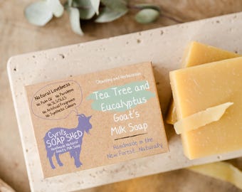May include: A close-up of handmade goat's milk soap bars and packaging. The packaging reads "Tea Tree and Eucalyptus Goat's Milk Soap" and "Cyril's Soap Shed". The soap is a light yellow color and is on a light-colored rectangular tray.