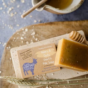 May include: A bar of Honey & Oats Goat's Milk Soap, with a wooden honey dipper and a small bowl of honey. The soap is light brown and sits on a rectangular stone. The soap is handmade and natural.