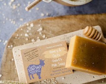 Può includere: Una saponetta al latte di capra al miele e avena, con un misurino di miele in legno e una piccola ciotola di miele. Il sapone è marrone chiaro e si trova su una pietra rettangolare.