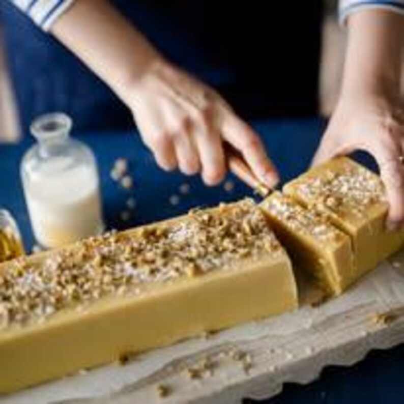 Pu&ograve; includere: Una persona taglia con un coltello una barra rettangolare di sapone fatto a mano. Il sapone &egrave; giallo chiaro e ricoperto di noci e semi marroni. Sullo sfondo ci sono una piccola bottiglia di vetro con liquido e un barattolo di olio.