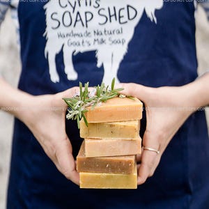 Pu&ograve; includere: Una pila di saponi artigianali al latte di capra in varie tonalit&agrave; di marrone e giallo, guarnita con rametti di rosmarino. I saponi sono tenuti davanti a un grembiule blu navy con il testo "Cyril's SOAP SHED Handmade Natural Goat's Milk Soap" e una silhouette di capra.