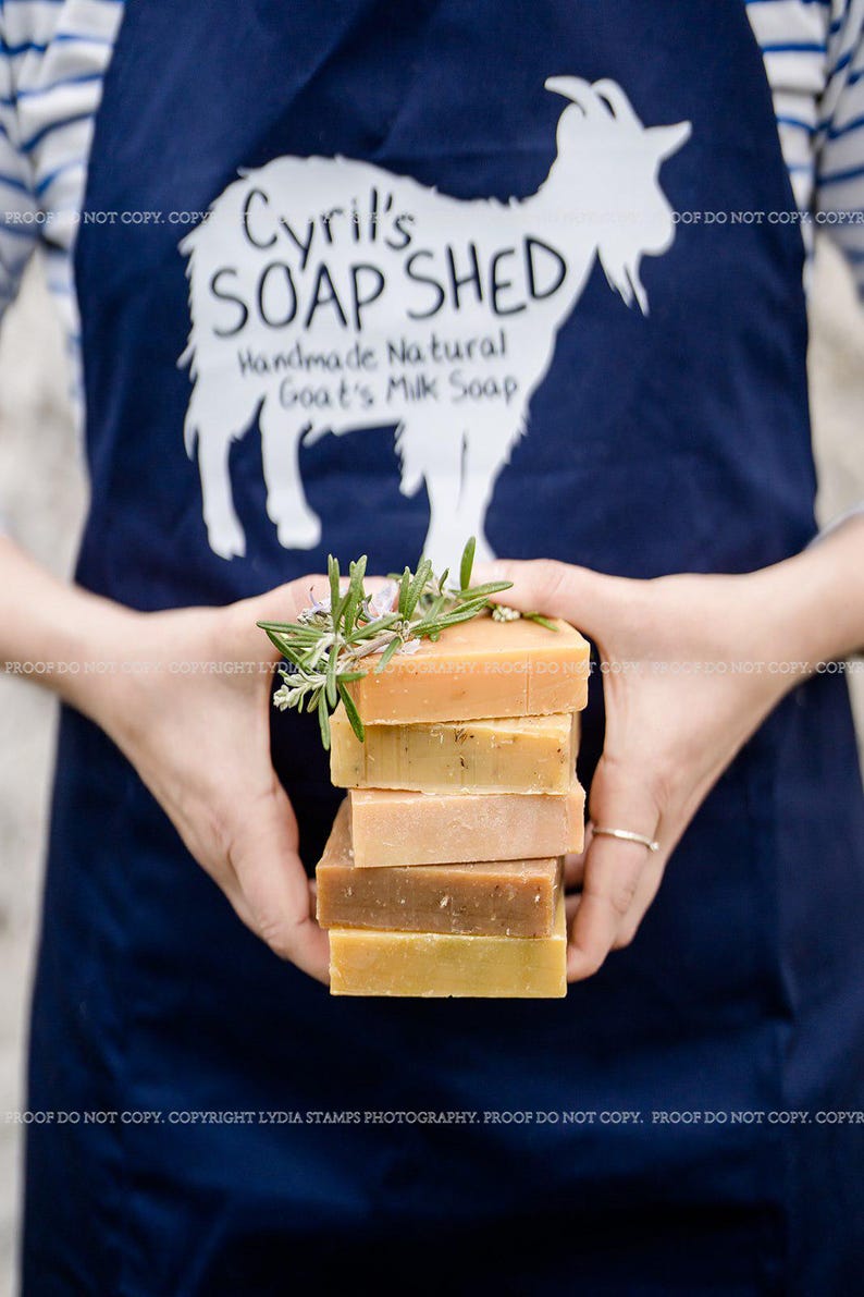 Pu&ograve; includere: Una pila di saponi artigianali al latte di capra in varie tonalit&agrave; di marrone e giallo, guarnita con rametti di rosmarino. I saponi sono tenuti davanti a un grembiule blu navy con il testo "Cyril's SOAP SHED Handmade Natural Goat's Milk Soap" e una silhouette di capra.