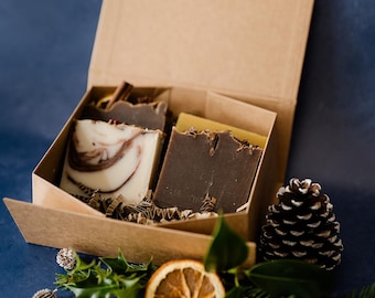 Può includere: Una confezione regalo con tre saponette fatte a mano. Una saponetta ha un motivo a spirale bianco e marrone, un'altra è marrone scuro e la terza è gialla. La scatola è decorata con pigne, fette d'arancia essiccate e verde.
