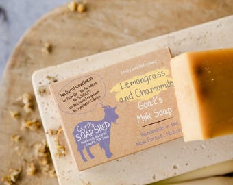 Può includere: Una saponetta al latte di capra e una scatola di sapone con la scritta "Lemongrass and Chamomile Goat's Milk Soap". La scatola riporta anche la scritta "Cyril's Soap Shed" e un'illustrazione di capra. Il sapone è su una superficie chiara.