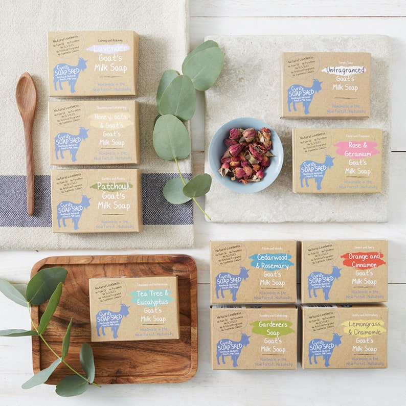 Pu&ograve; includere: Un assortimento di scatole di sapone al latte di capra in varie fragranze, tra cui Lavanda, Miele e Avena, e Tea Tree Eucalipto. Le scatole marroni hanno etichette blu e bianche. L'immagine include foglie di eucalipto e un cucchiaio di legno.