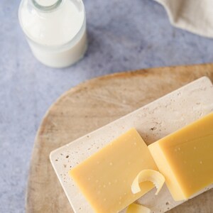 Pu&ograve; includere: Una natura morta con un blocco di formaggio giallo su un tagliere rettangolare in pietra, con scaglie di formaggio. Una piccola bottiglia di vetro con un liquido bianco &egrave; vicina su un tagliere di legno. Un panno beige &egrave; sullo sfondo.