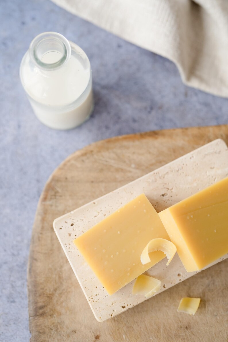 Pu&ograve; includere: Una natura morta con un blocco di formaggio giallo su un tagliere rettangolare in pietra, con scaglie di formaggio. Una piccola bottiglia di vetro con un liquido bianco &egrave; vicina su un tagliere di legno. Un panno beige &egrave; sullo sfondo.
