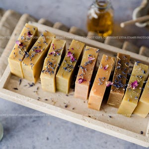 Pu&ograve; includere: Una collezione di saponi fatti a mano in varie tonalit&agrave; di giallo e marrone, decorati con fiori secchi. I saponi sono disposti su un vassoio rettangolare in pietra. Una bottiglia di olio e una ciotola di miele sono sullo sfondo.