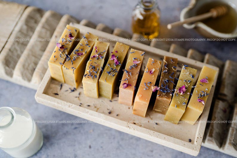 Pu&ograve; includere: Una collezione di saponi fatti a mano in varie tonalit&agrave; di giallo e marrone, decorati con fiori secchi. I saponi sono disposti su un vassoio rettangolare in pietra. Una bottiglia di olio e una ciotola di miele sono sullo sfondo.