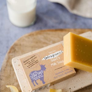 Pu&ograve; includere: Una saponetta al latte di capra non profumata di colore giallo chiaro &egrave; appoggiata su un portasapone in pietra. La confezione recita "Sapone al latte di capra non profumato". Sullo sfondo, una piccola bottiglia di latte.