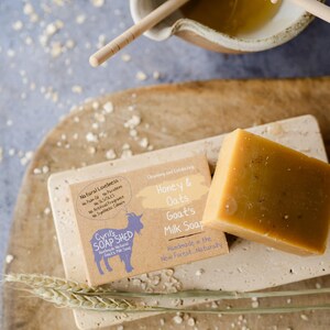 May include: A close-up of handmade goat's milk soap with honey and oats. The soap is a light brown color and is labeled "Honey & Oats Goat's Milk Soap." The image also shows a wooden bowl with honey and a honey dipper.