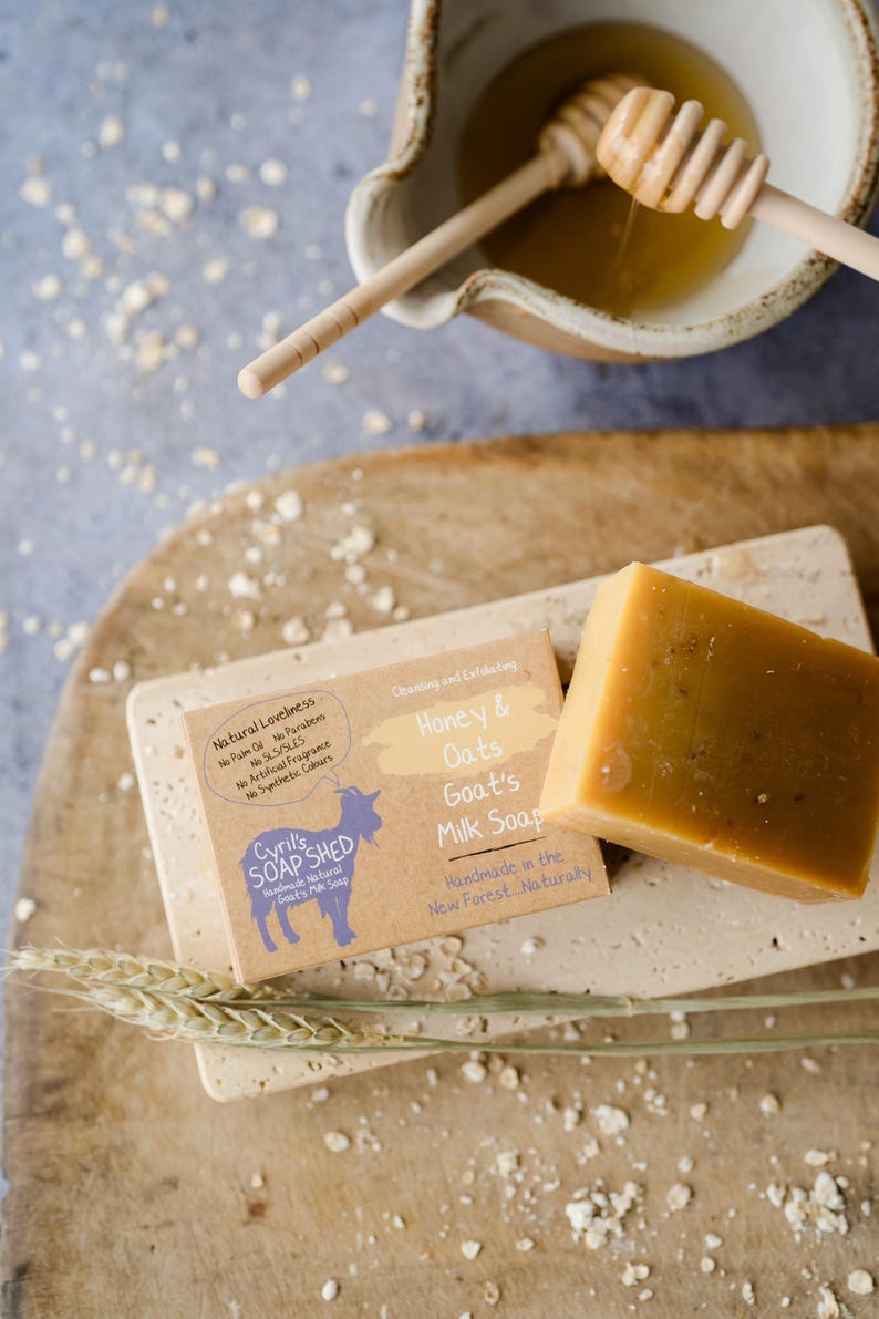 May include: A close-up of handmade goat's milk soap with honey and oats. The soap is a light brown color and is labeled "Honey & Oats Goat's Milk Soap." The image also shows a wooden bowl with honey and a honey dipper.
