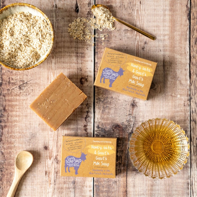 May include: Overhead shot of natural soap bars, ingredients, and packaging. The soap bars are light brown, with packaging that reads "Honey, oats, & Goat's Milk Soap." A bowl of oats, a spoon, and a small glass dish are also present.
