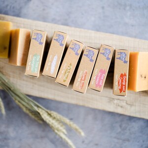 May include: An overhead shot of handmade soaps and their packaging. The soaps are various shades of yellow and orange. The packaging is brown with a goat logo and text indicating the scent, such as "Tea Tree and Eucalyptus".