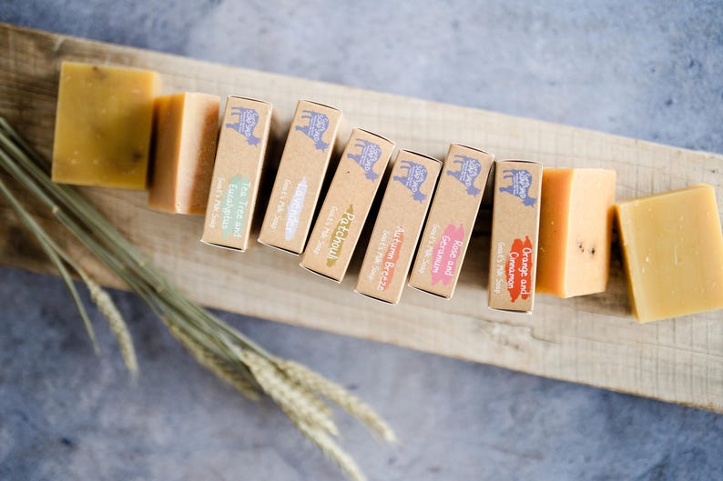 May include: An overhead shot of handmade soaps and their packaging. The soaps are various shades of yellow and orange. The packaging is brown with a goat logo and text indicating the scent, such as "Tea Tree and Eucalyptus".