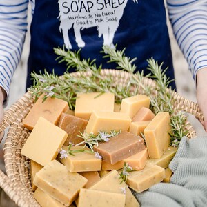 Pu&ograve; includere: Un cesto di vimini pieno di saponette artigianali al latte di capra in varie tonalit&agrave; di giallo e marrone. Il cesto &egrave; decorato con rametti di rosmarino e piccoli fiori viola. Il grembiule sullo sfondo recita "Cyril's Soap Shed".