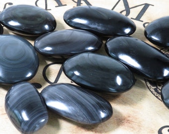 Op de afbeelding: Een verzameling gladde, ovale zwarte obsidiaanstenen met een glanzende afwerking. De stenen hebben een draaiende, gebandeerde structuur.