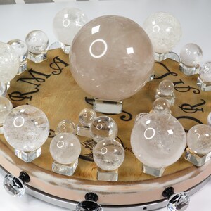 May include: A collection of clear quartz crystal spheres of various sizes displayed on a wooden base with decorative knobs. The largest sphere is a light pink color.
