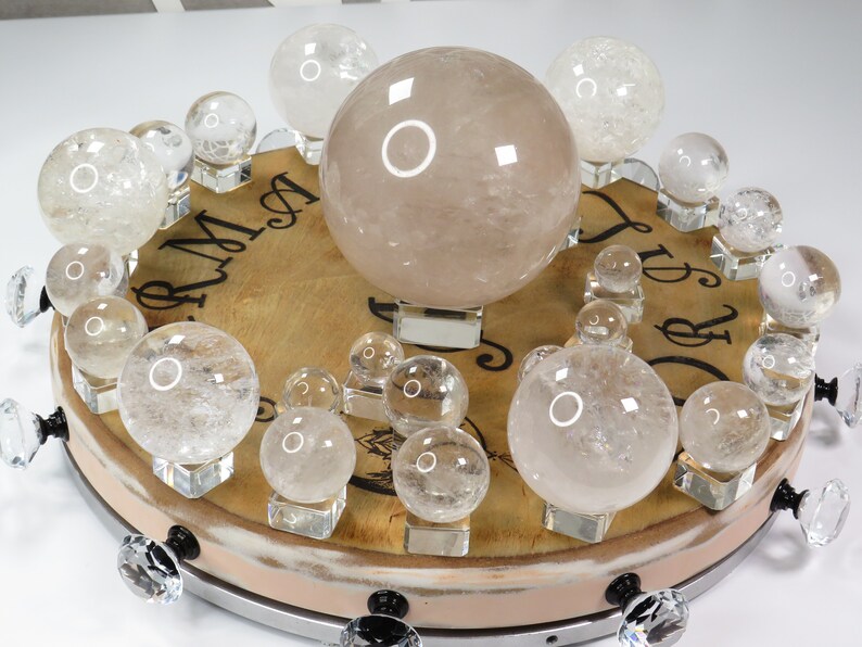May include: A collection of clear quartz crystal spheres of various sizes displayed on a wooden base with decorative knobs. The largest sphere is a light pink color.
