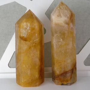 May include: Two golden citrine crystal points, one slightly larger than the other, are displayed on a white surface. The crystals are clear with a golden hue and have a natural, faceted shape.