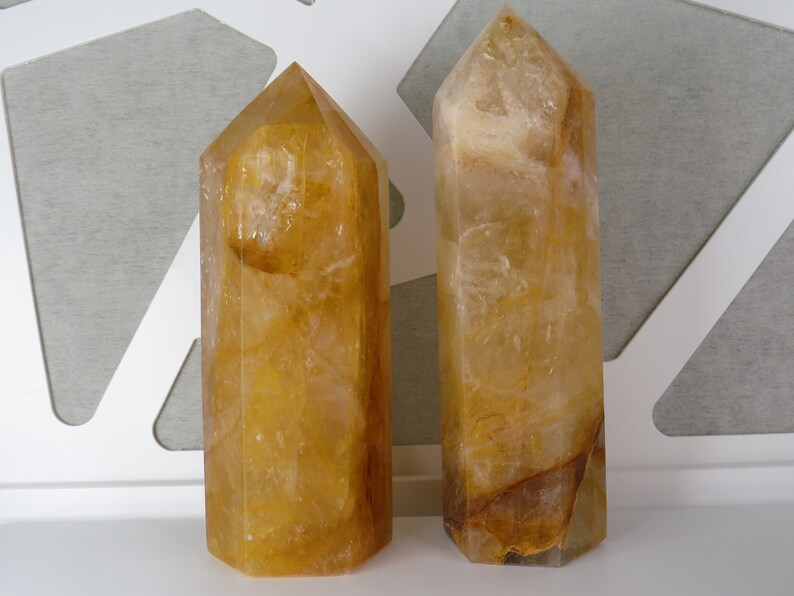 May include: Two golden citrine crystal points, one slightly larger than the other, are displayed on a white surface. The crystals are clear with a golden hue and have a natural, faceted shape.