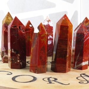 May include: A group of red, orange, and white jasper crystal points arranged on a wooden surface with the word "CORAL" printed on it.