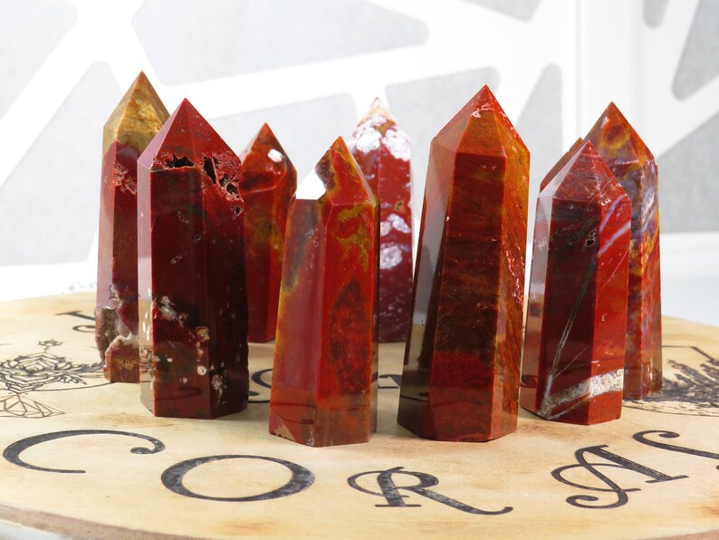 May include: A group of red, orange, and white jasper crystal points arranged on a wooden surface with the word "CORAL" printed on it.