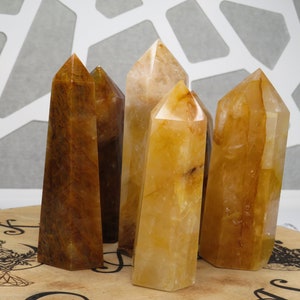 May include: Five golden citrine crystal points, some with a brown hue, arranged on a wooden surface with a decorative design.