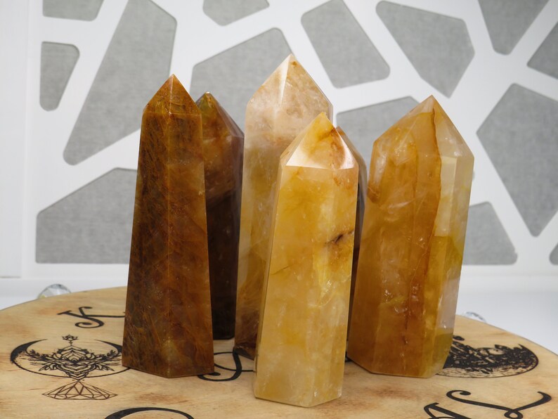 May include: Five golden citrine crystal points, some with a brown hue, arranged on a wooden surface with a decorative design.