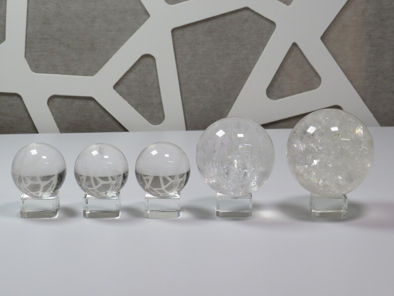 May include: Five clear quartz crystal spheres of varying sizes, displayed on small square bases. The spheres are smooth and translucent, with a slight sparkle.