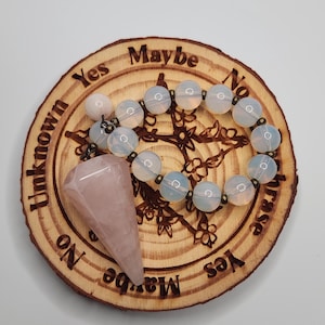 Könnte beinhalten: Ein hellrosa facettiertes Kristallpendel liegt auf einem runden Holzbrett, in das die Wörter Yes, Maybe und No eingraviert sind. Ein Armband aus durchscheinenden weißen Perlen mit kleinen metallischen Zwischenstücken liegt auf der Holzfläche.