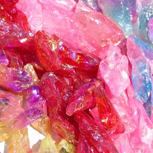 May include: A collection of iridescent crystal points in various shades of pink, red, purple, and blue. The crystals are arranged in a random pattern on a white surface.