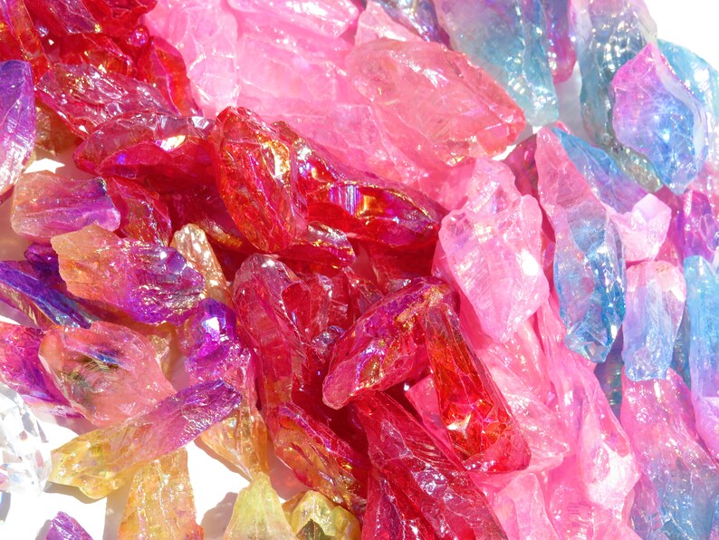 May include: A collection of iridescent crystal points in various shades of pink, red, purple, and blue. The crystals are arranged in a random pattern on a white surface.