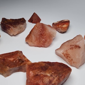 May include: A collection of eight rough, natural, pink and brown carnelian crystals. The crystals are various sizes and shapes.