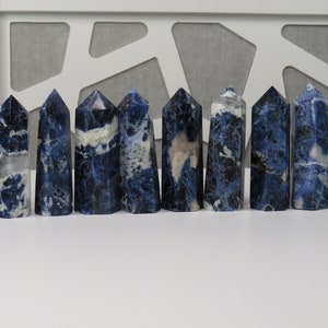 May include: A collection of ten polished sodalite crystal towers arranged in a horizontal row. Each tower features a hexagonal base and a pointed top, displaying a mix of deep blue, navy, and white marbled patterns with unique mineral inclusions.